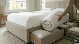 A photorealistic, high-detail illustration showing a close-up of a light beige divan double bed. A person is neatly placing a folded, heavy-duty white winter duvet and two substantial pillows into the deep, opened side-access storage drawer to show seasonal organisation.
