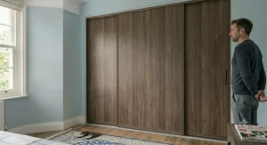 A closed three-door sliding wardrobe with a premium dark wood-grain effect finish, seamlessly integrated into a modern bedroom interior.